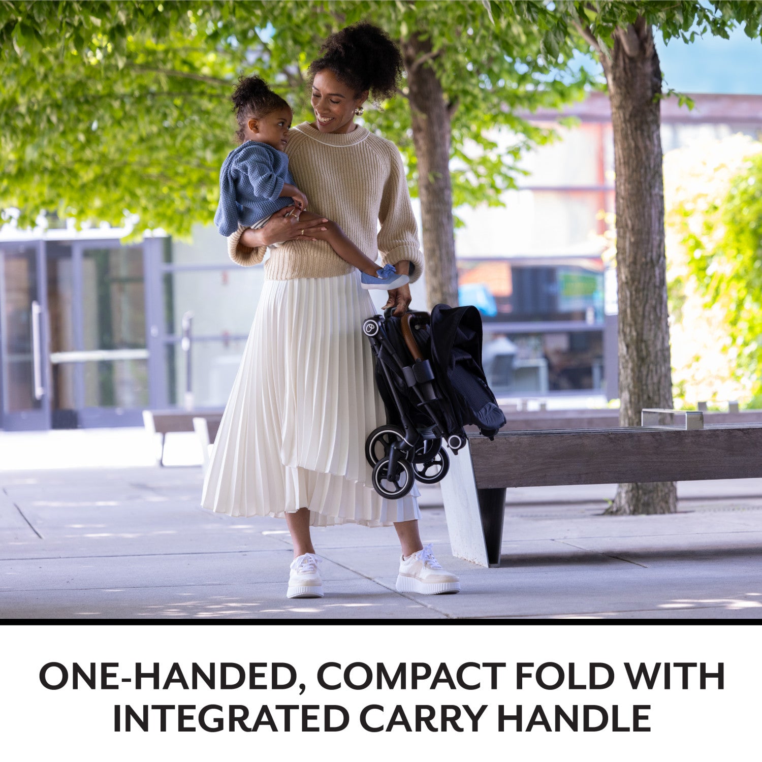 You are a happy parent, holding a baby and a folded stroller in a park.