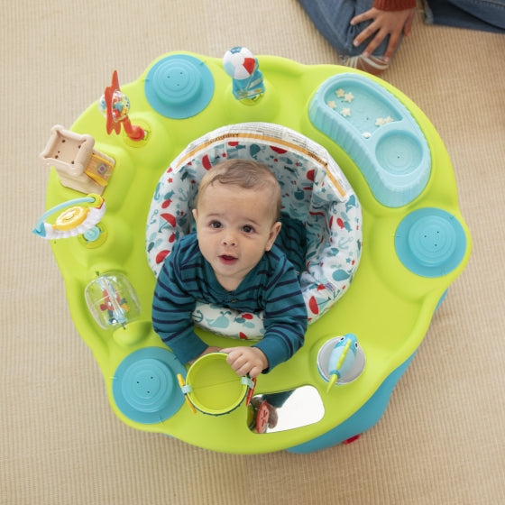A top-down view of a baby happily seated in a light green and blue activity center, surrounded by various toys.