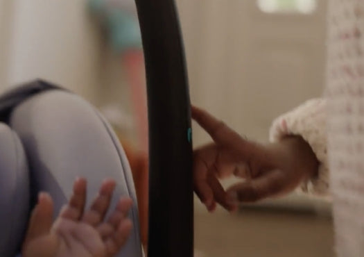 Hand pressing a button on the handle of a car seat, with a baby's hand visible in the seat.