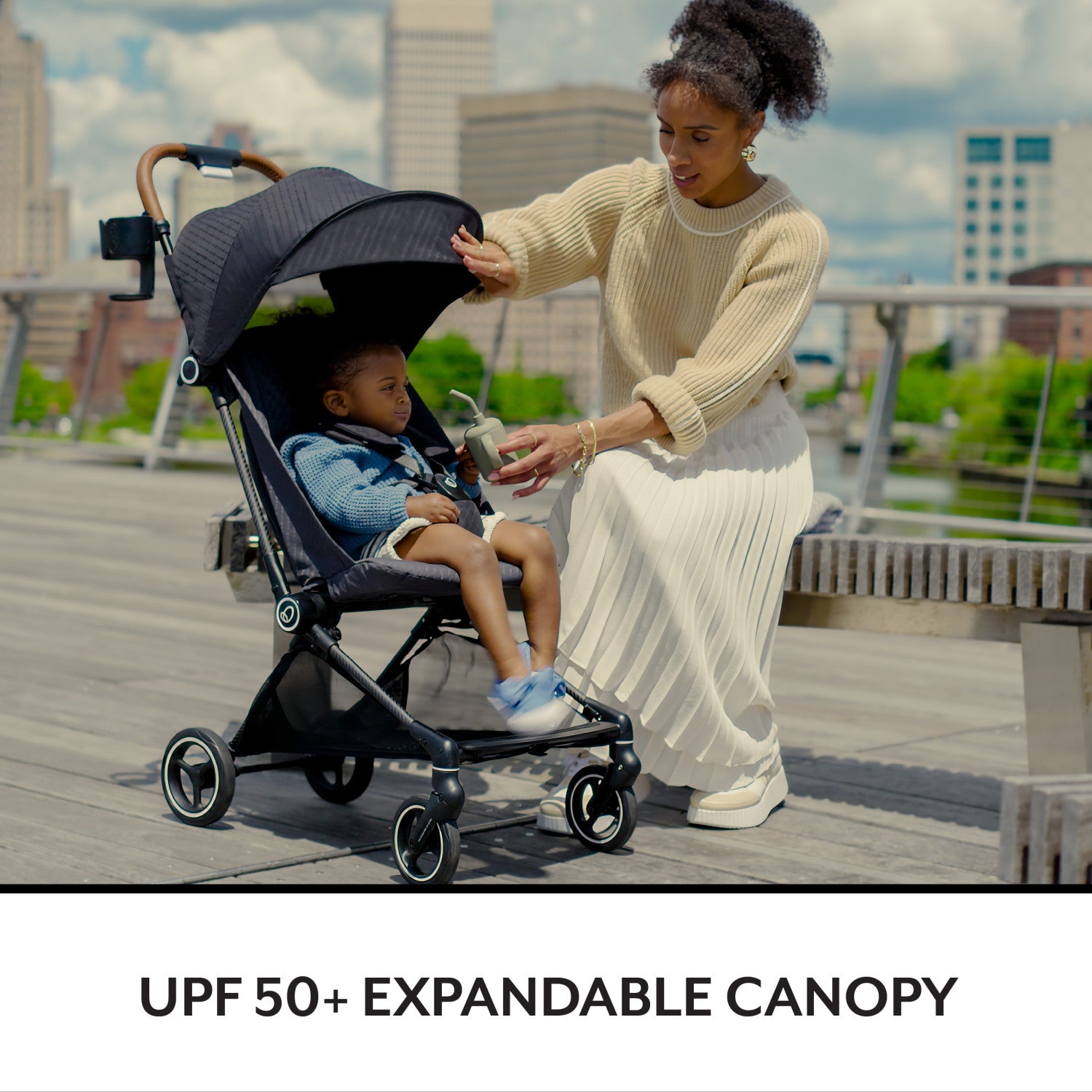 A loving mother on a city bridge adjusts the canopy and gives her child a drink in a modern stroller.