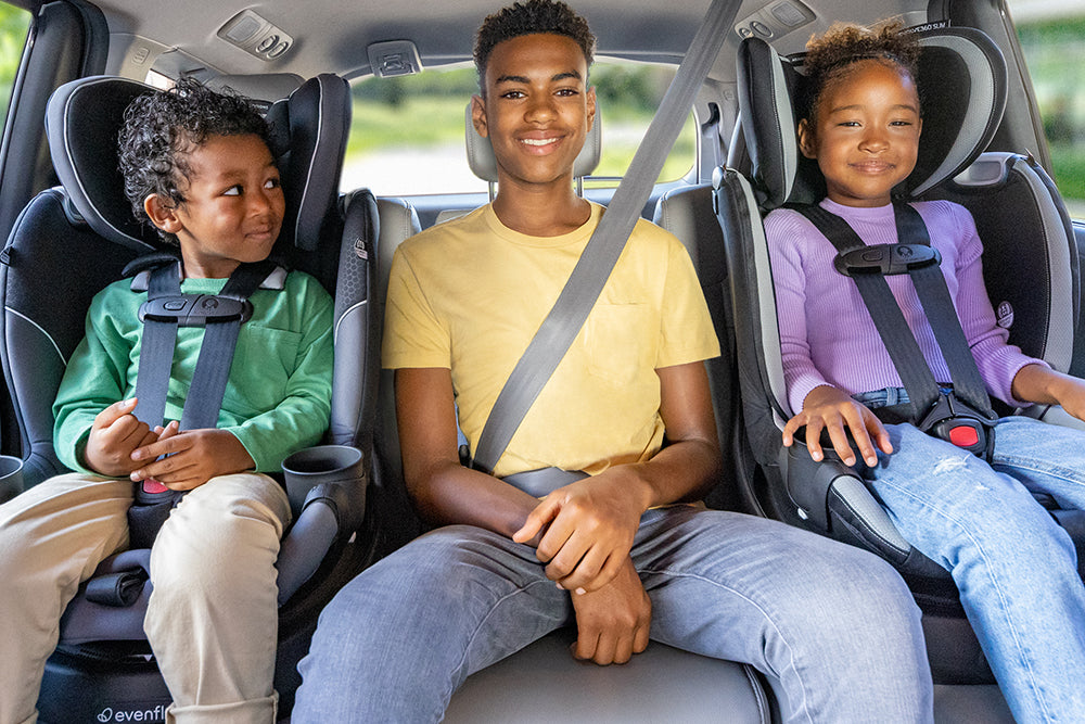 Three children, two in car seats and one using a seat belt, are sitting in the back seat of a car. They are all smiling.