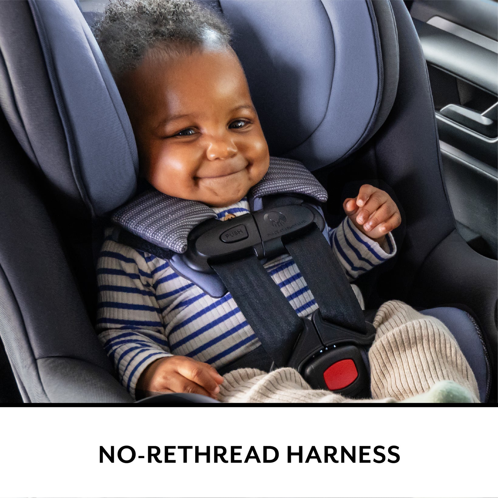 A smiling baby safely secured in a gray car seat, wearing a striped long-sleeved shirt.