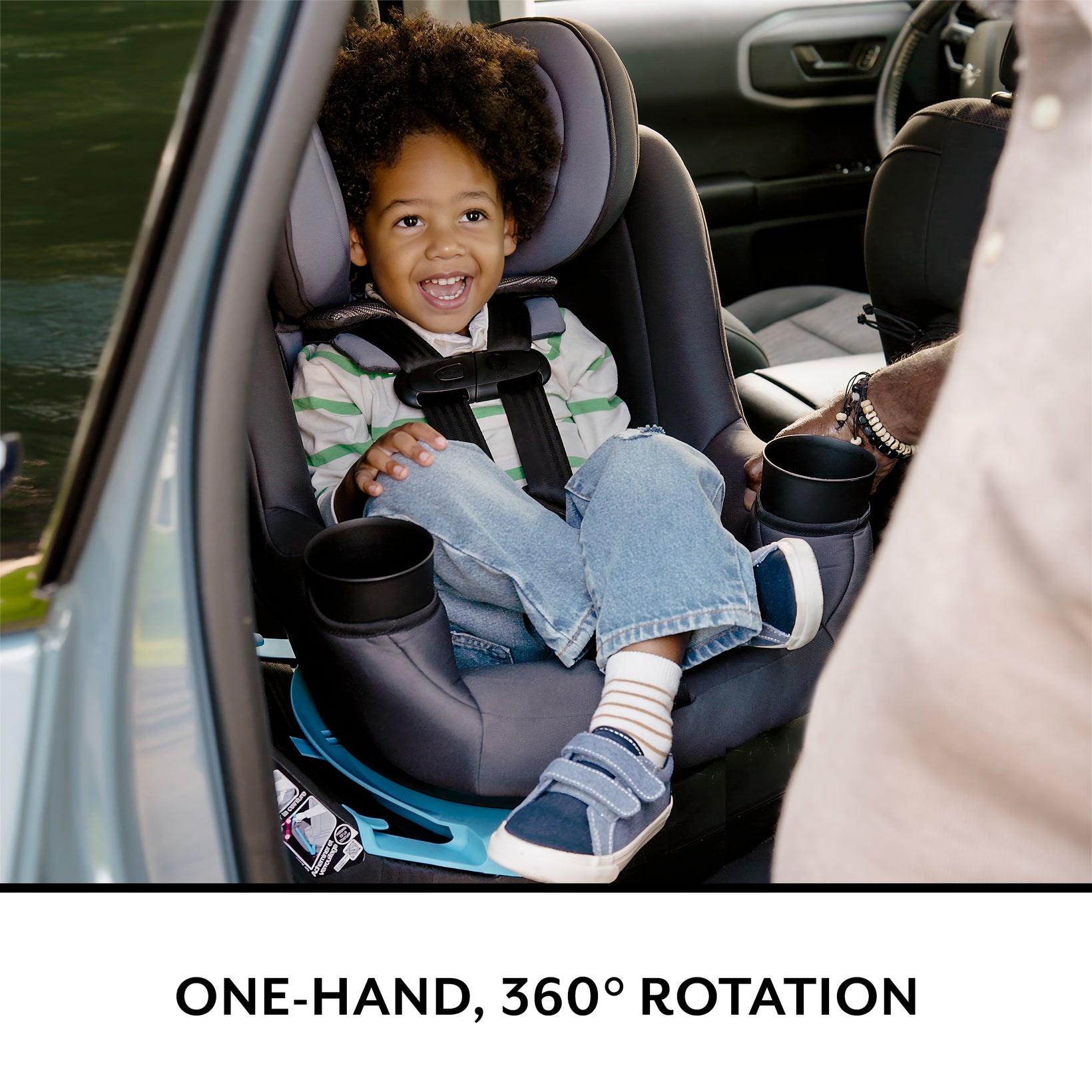A smiling young child safely seated in a gray car seat with two cup holders.