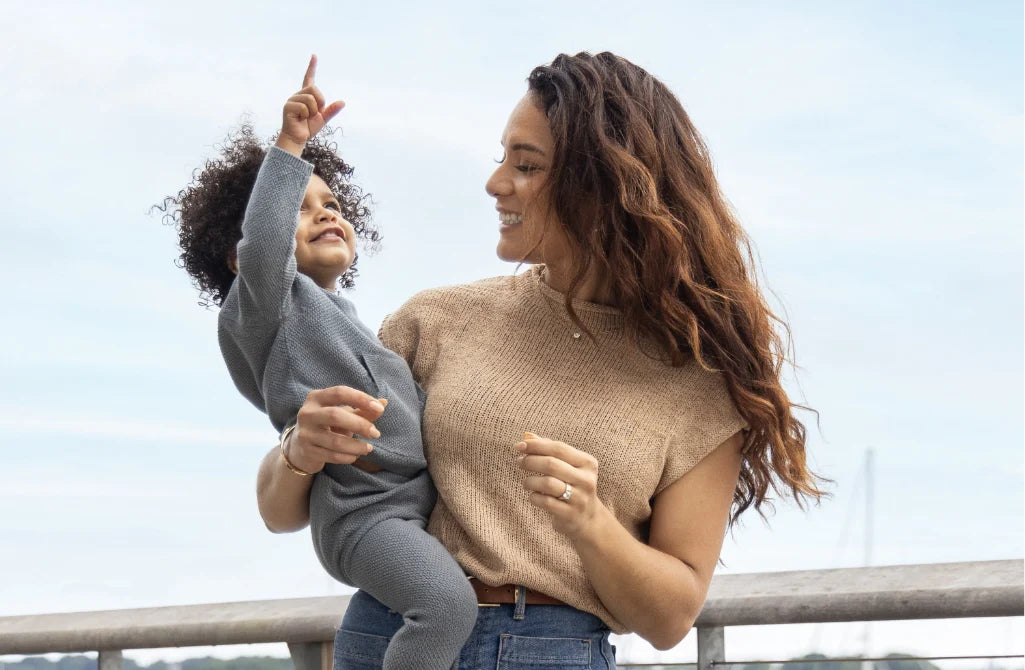 Woman holding a child and pointing, both smiling outdoors.