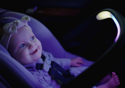 A happy baby with a headband, secured in a car seat with a glowing light strip on the handle.