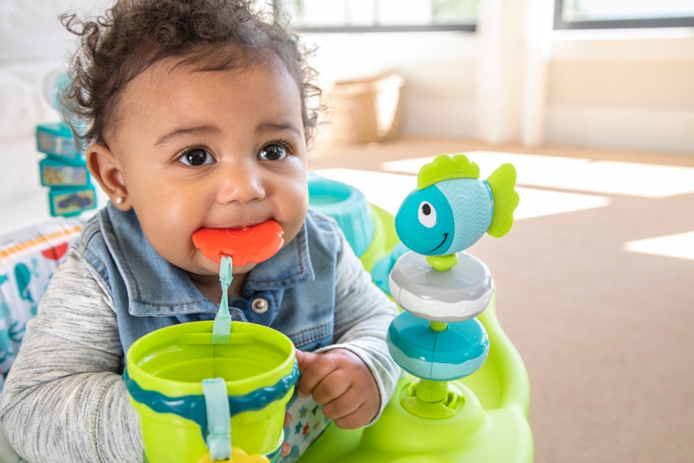 A baby in an activity center chews on a teether toy attached to a cup, next to a spinning fish toy.