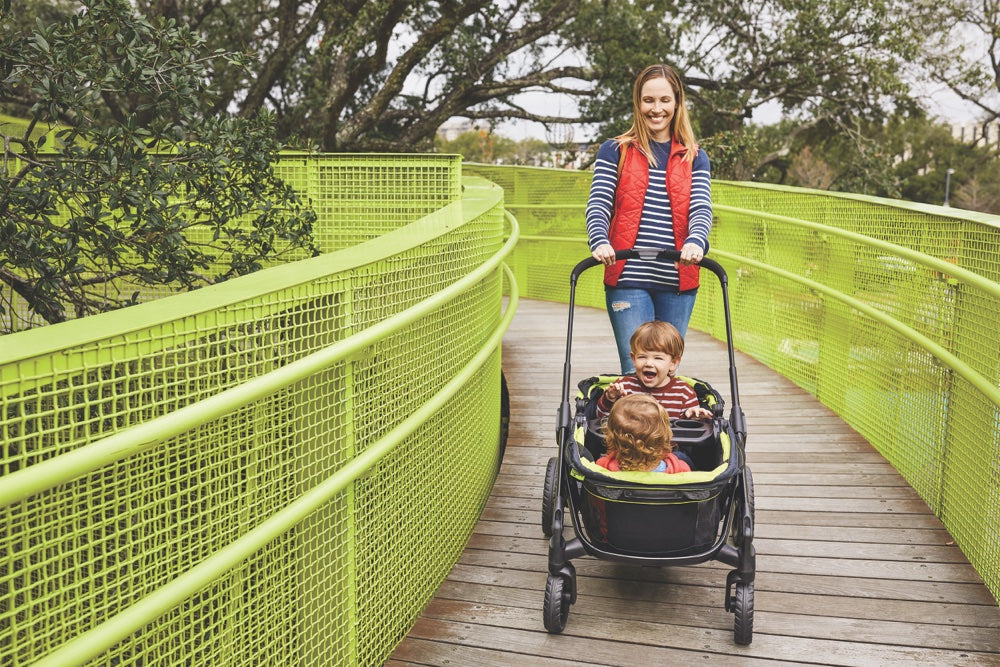 A smiling woman pushes a black side-by-side double stroller with two happy children along a walkway with bright green railings.