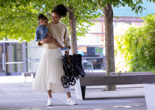 You are a happy parent, holding a baby and a folded stroller in a park.
