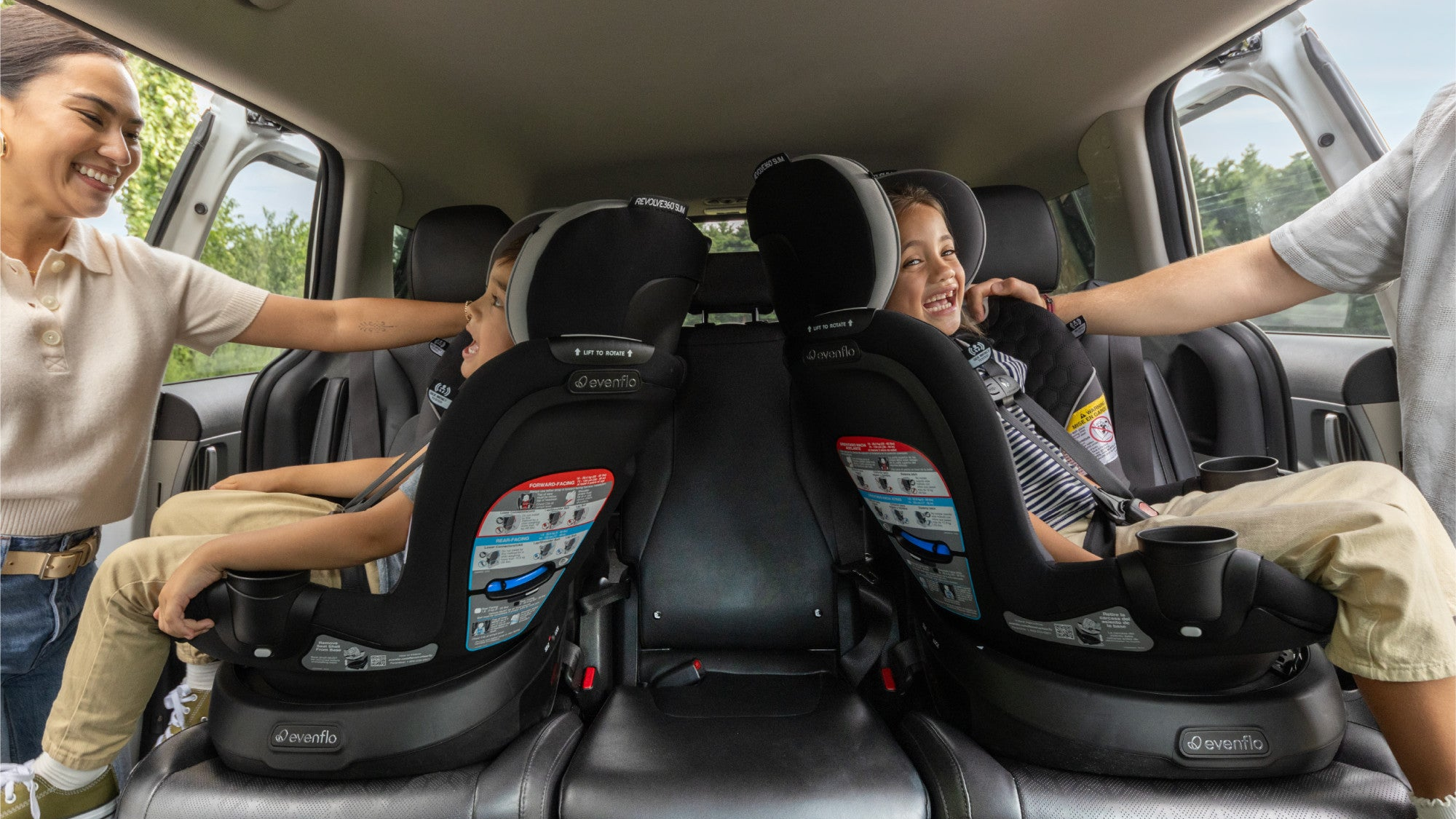 Two children in car seats with a woman and man assisting them in a vehicle.