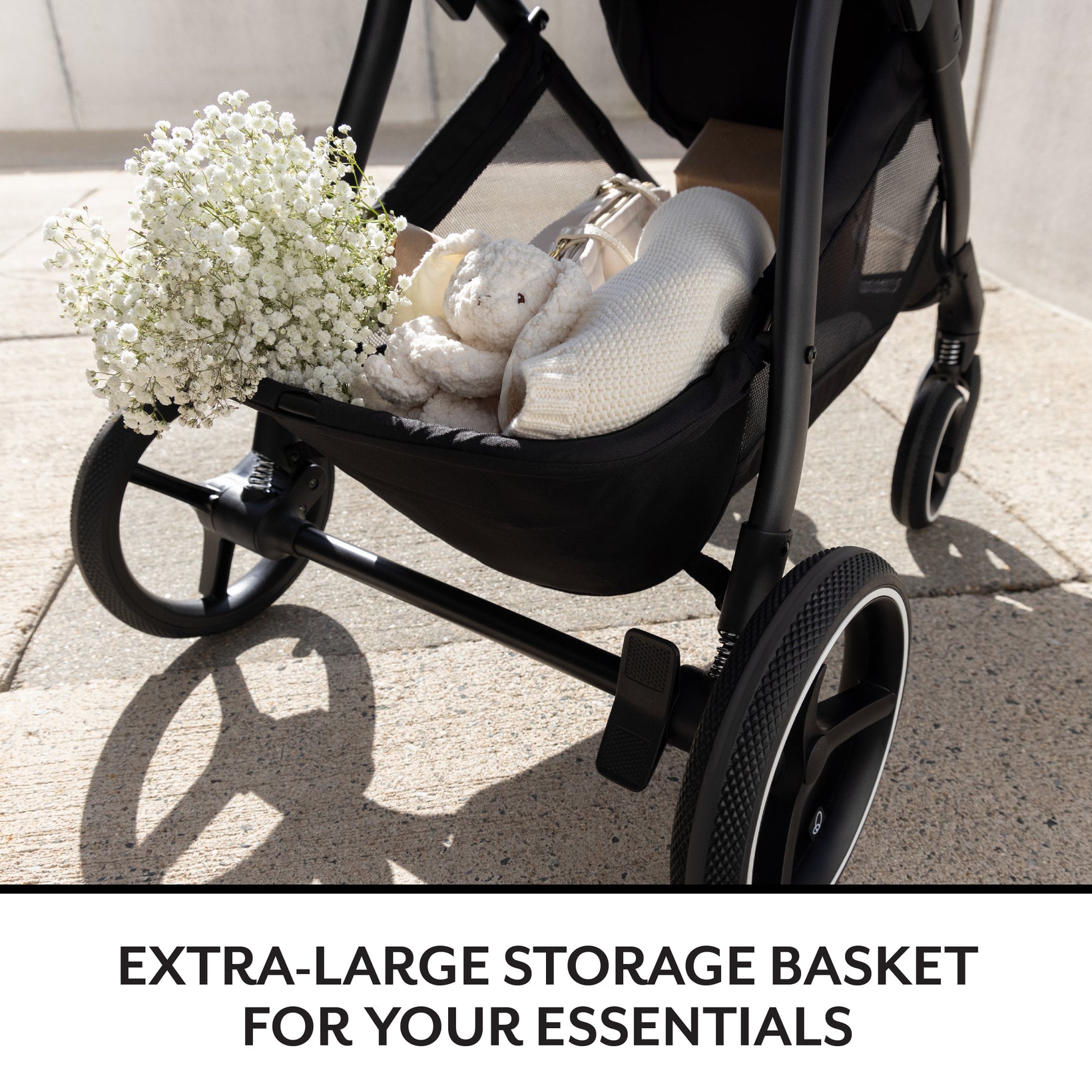 Black stroller with an extra-large storage basket containing a teddy bear, flowers, and a blanket on a concrete surface.